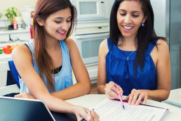 Two Individuals Are Collaborating At A Kitchen Table With A Laptop Open And Documents In Front Of Them. They Are Smiling As One Person Points At A Paper While The Other Looks On. They Appear Engaged In A Productive Discussion.