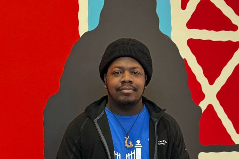 Young Man Wearing A Beanie, A Blue College Possible Shirt And A Black Jacket Standing In Front Of A Brightly Colored Wall.