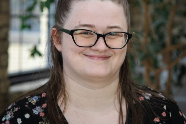 Portrait Of A Smiling Person Wearing Glasses And A Floral Shirt, Standing In A Room With Green Plants In The Background.