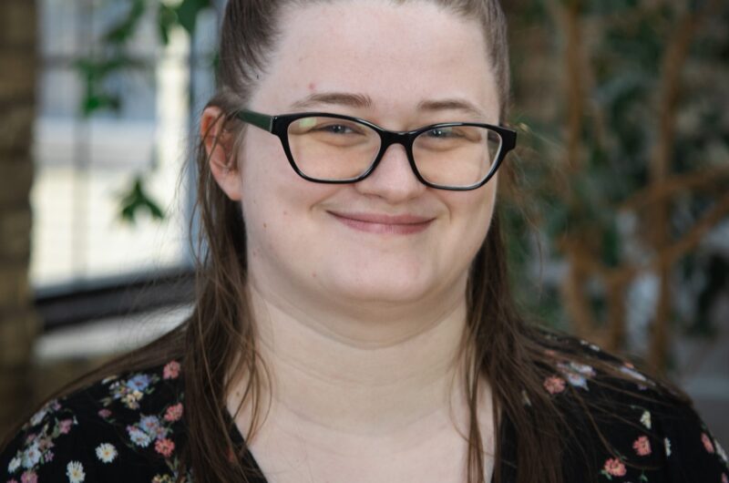 Portrait Of A Smiling Person Wearing Glasses And A Floral Shirt, Standing In A Room With Green Plants In The Background.