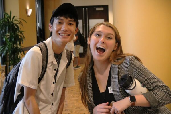 Two Individuals Smiling Happily, One Wearing A Cap And Carrying A Backpack, And The Other Dressed In A Blazer Holding A White Water Bottle And A Clipboard.