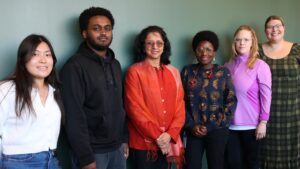 Group of six diverse individuals smiling and standing in a row against a green wall.