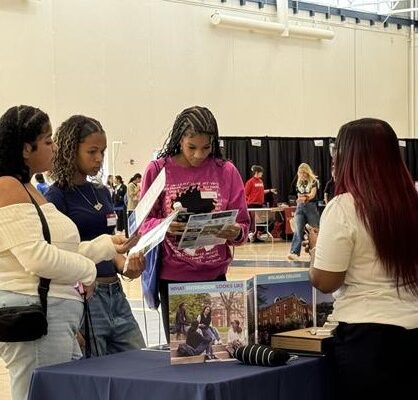 Three Individuals Are Attentively Reading Brochures At A College Fair Booth Staffed By A Representative. The Booth Features Various Informational Materials And A Banner Of Rutgers University.