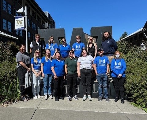 People stand in two rows in front of the letter W with greenery on either side and a W on a flag in the background.