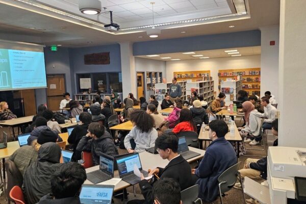 Group Of About Students In A Library Seated At Tables With Laptops, All Facing A Projector Where Information About FAFSA Is Displayed.