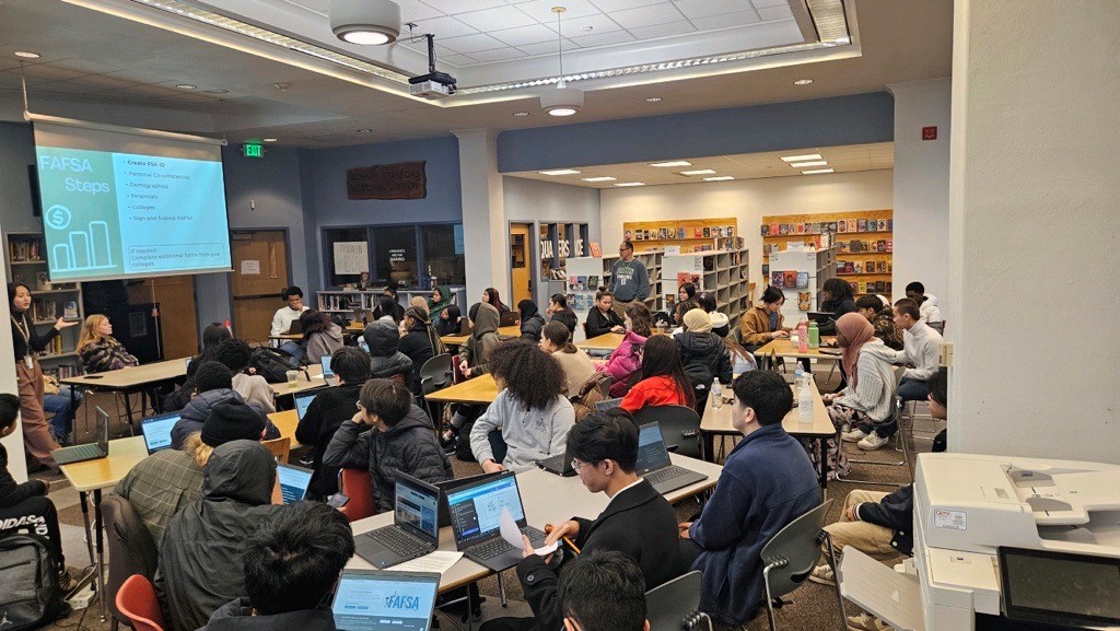 Group of about students in a library seated at tables with laptops, all facing a projector where information about FAFSA is displayed.