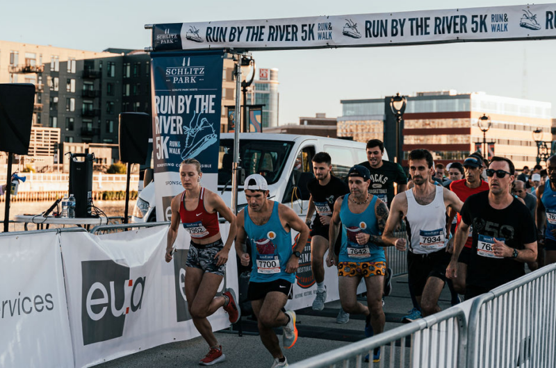 A Group Of People Dressed In Brightly Colored Athletic Clothing Running Past The Run By The River Schlitz Park Starting Line.