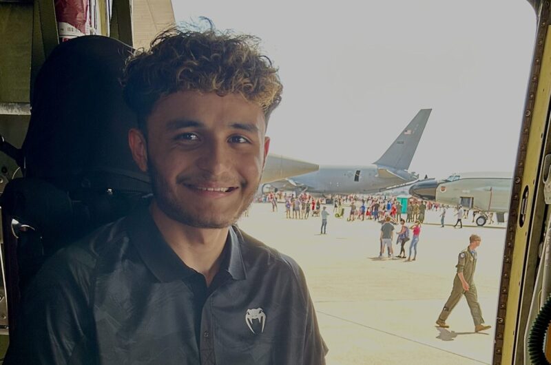 Daniel Smiling At The Camera With His Thumbs Up Sitting Inside An Aircraft With The Door Open Behind Him Revealing Other Planes And People On The Tarmac.