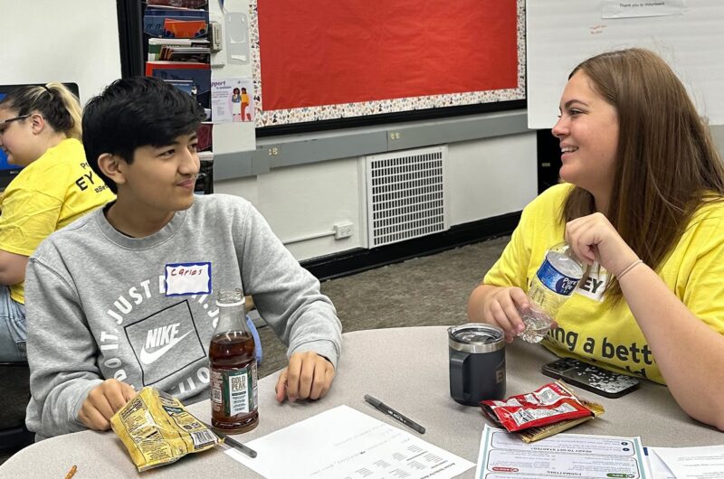 A Student And A Volunteer In A Yellow Shirt Talk At A Classroom Table With Papers On It.