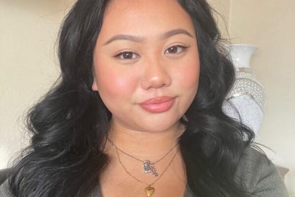 Portrait Of A Person With Dark Wavy Hair And A Beige Top, Wearing A Necklace, Sitting In A Well-lit Room.