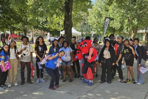 A Large Group Of Students And Three Coaches In Blue Polos Pose For A Photo With University Of Illinois-Chicago’s Dragon Mascot On A College Campus.