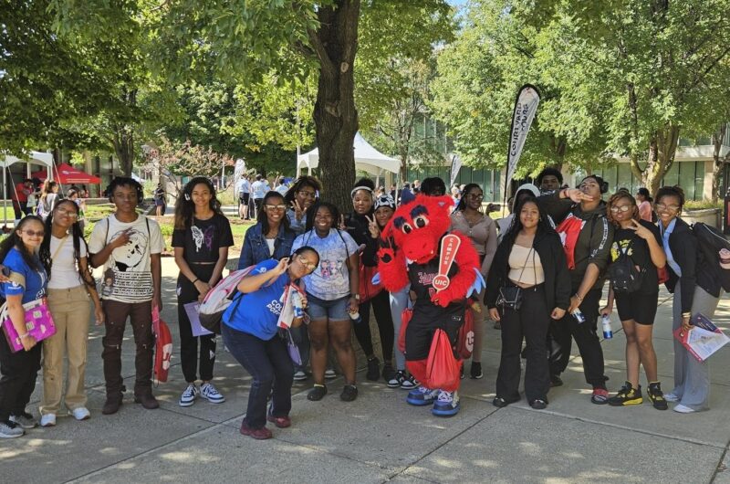 A Large Group Of Students And Three Coaches In Blue Polos Pose For A Photo With University Of Illinois-Chicago’s Dragon Mascot On A College Campus.