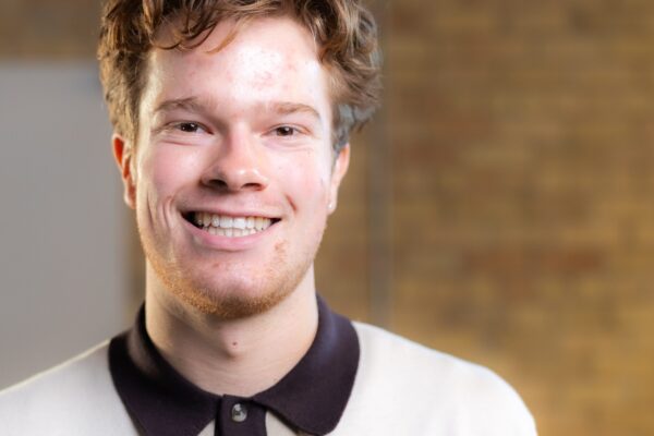 Person Smiling At The Camera In A Collared Shirt, Standing In Front Of A Brick Wall.