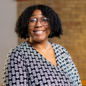 Portrait Of A Smiling Person With Glasses, Wearing A Floral Top, Against A Blurred Brick Wall Background.