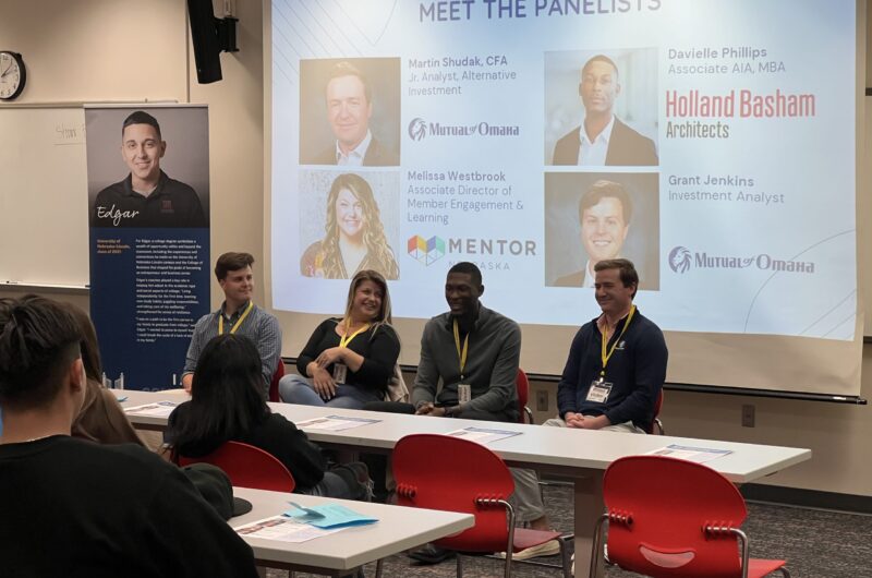 Photo Of Four Panelists Seated In Front Of A Projection Screen Showing Their Names, Photos And Titles.