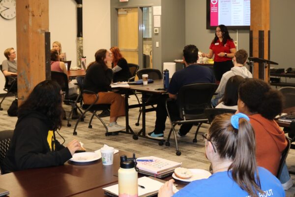 A Person Presenting To A Group Of Attentive Listeners In A Seminar Room At A College Event. The Presentation Displayed On The Screen Reads "Meet The Colleges.