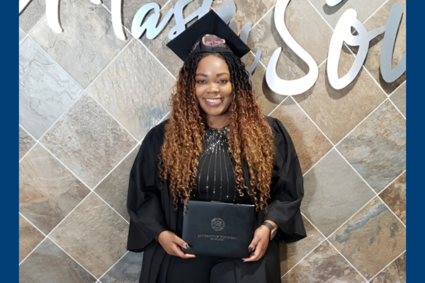 Graduate With Long Brown Hair Dressed In A Cap And Gown Standing In Front Of A Tiled Wall Holding Her Diploma