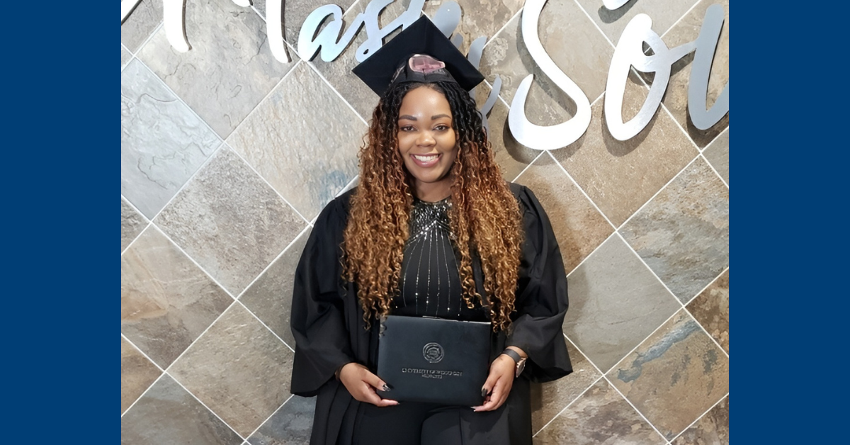 Graduate with long brown hair dressed in a cap and gown standing in front of a tiled wall holding her diploma