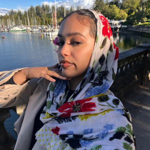 Close-up Of A Person Wearing A Floral Scarf, Posing With Their Hand On Their Chin, With A Backdrop Of A Sunny Marina Full Of Boats.