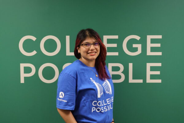 Headshot Photo Of Liz Smiling, Wearing Glasses And A Blue College Possible T-shirt With A Green Background Behind Her With “College Possible” Written On It.