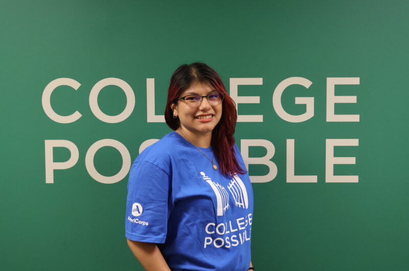 Headshot Photo Of Liz Smiling, Wearing Glasses And A Blue College Possible T-shirt With A Green Background Behind Her With “College Possible” Written On It.