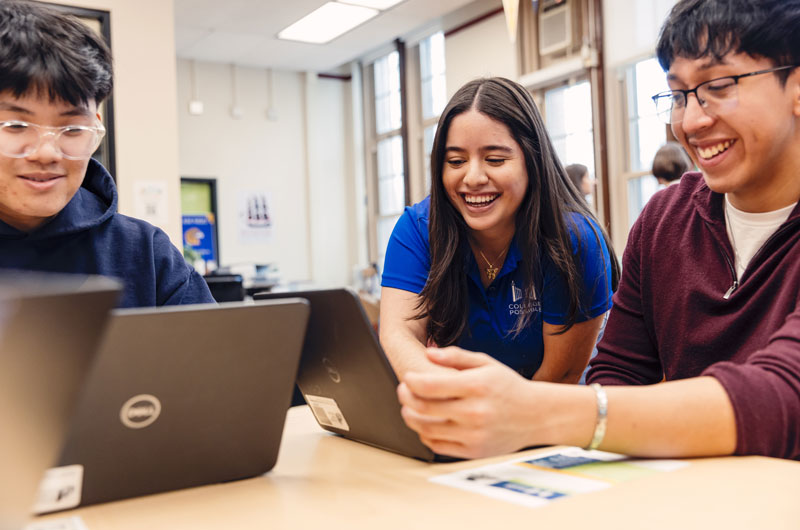 Two students sit at a table using laptops smiling. A College Possible coach helps one of the students with a task on the laptop.