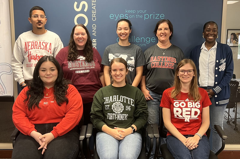Five leadership team members stand next to each other smiling with three leadership team members seated in a row in front of them smiling at the camera.