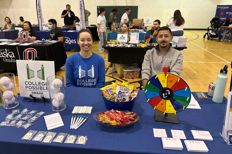 Advising specialists Dorothy (left) and Edgar (right) smiling and sitting at a table with a blue table cloth on it, with College Possible promotional materials and snacks spread out.