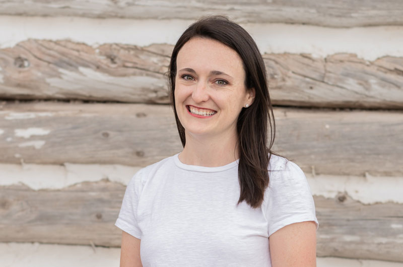 A woman wearing a white shirt stands in front of a wooden wall.