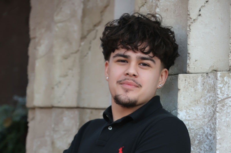 Hector stands smiling with his back against a white stone wall with his arms crossed in front of his chest.