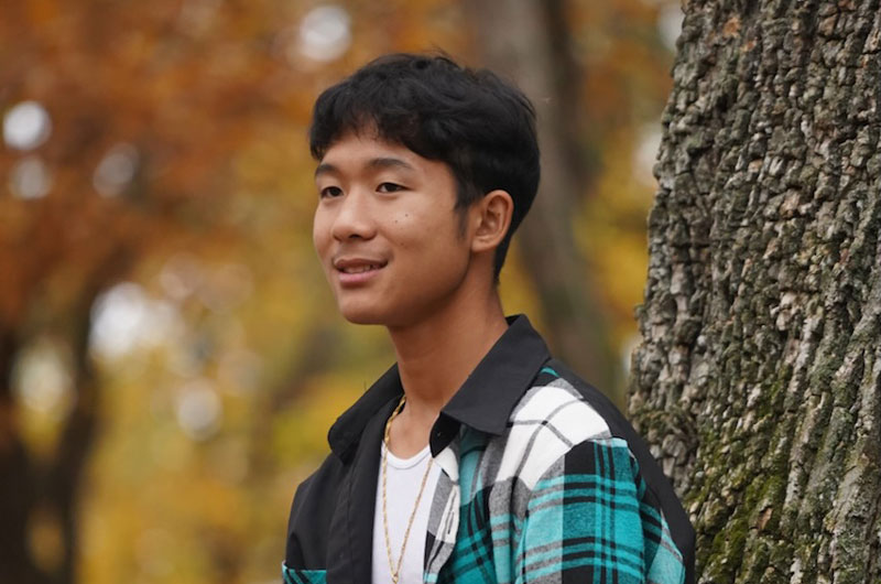 Saw Htoo stands with his back leaning against a tree, wearing a white shirt with a blue, black, and white flannel over top, smiling and looking into the distance.
