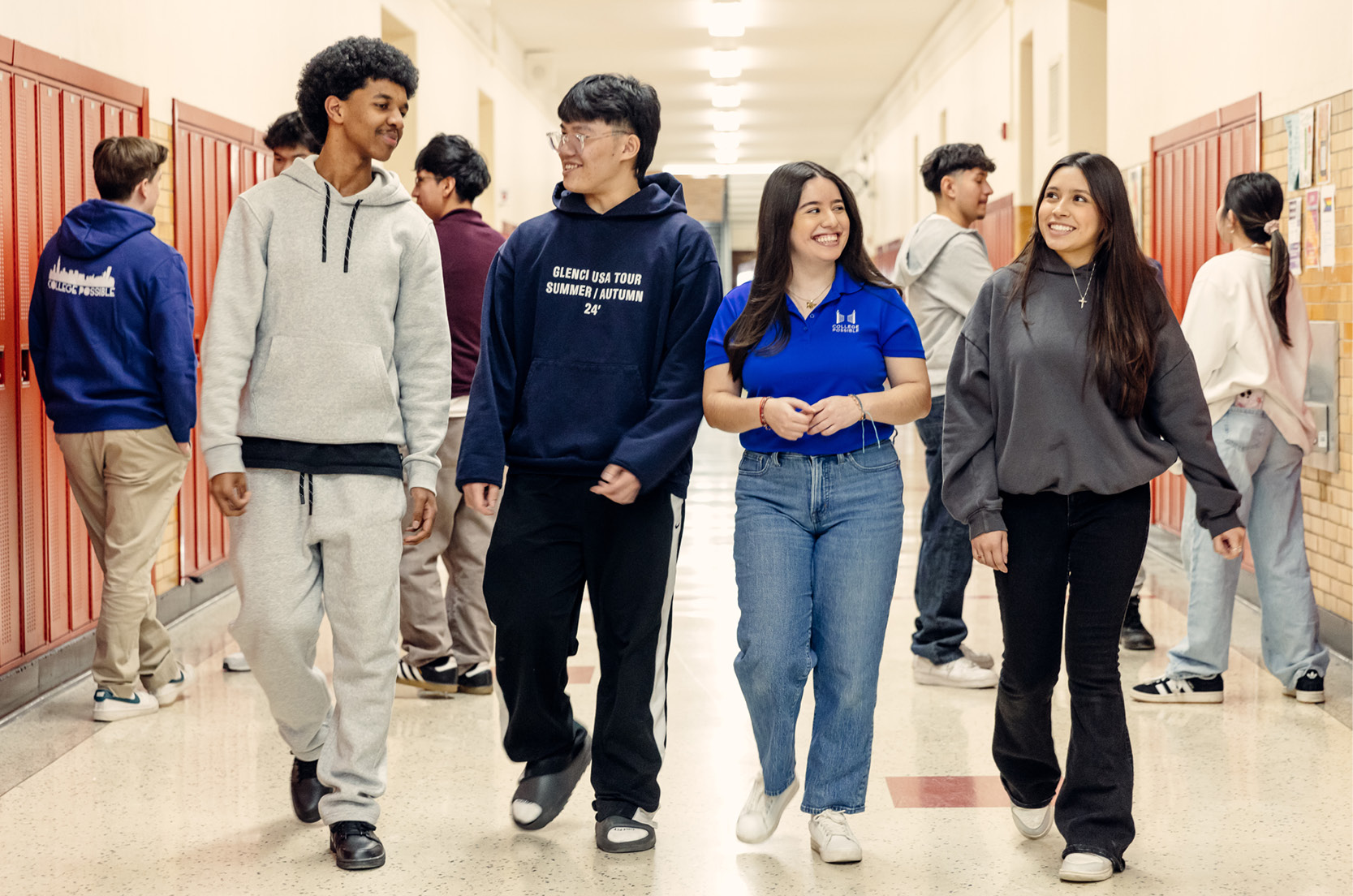 A group of three students and a coach walk side by side through a high school, with lockers on either side