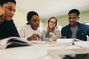 Four young adults sit together at a table, studying and discussing with notebooks and textbooks open in front of them.