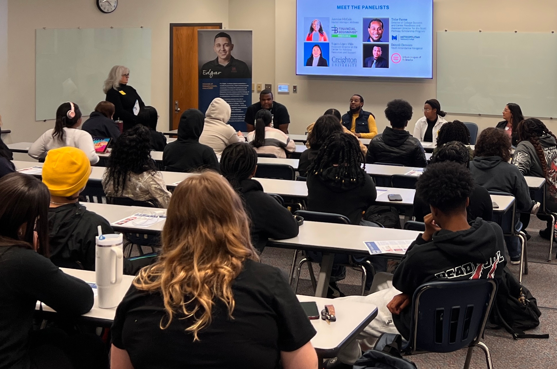 Panelists sit at the front of the room facing students with a tv screen behind them displaying their photos and job titles. Several rows of students are seated facing the panelists.