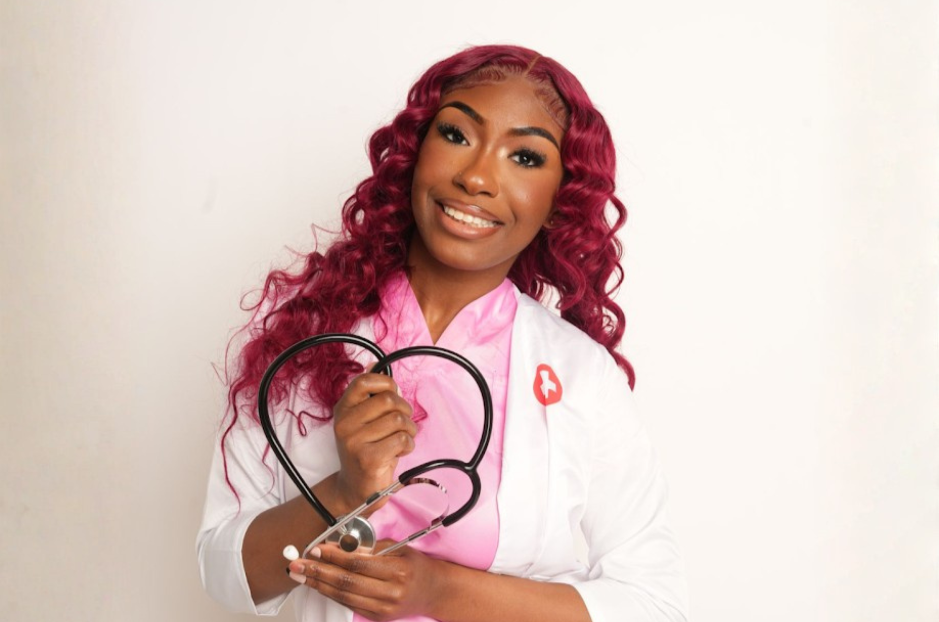 A student wearing pink scrubs and a lab coat poses smiling with a stethoscope in a heart shape. 