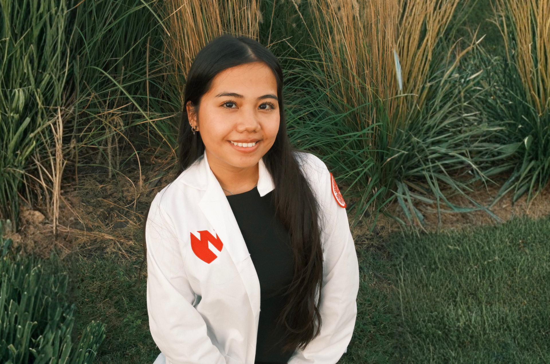 Photo of Nye smiling with a grassy background behind her. She wears a white UNMC coat over a black top.