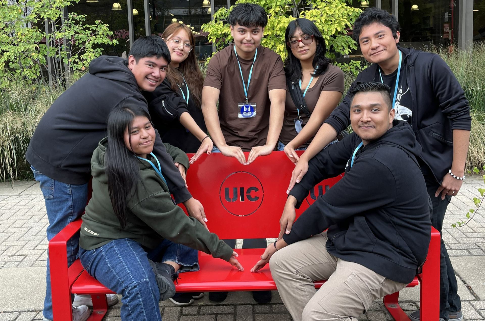 A group of students pose outside on a college campus, forming a heart with their hands around the UIC logo on a bench.