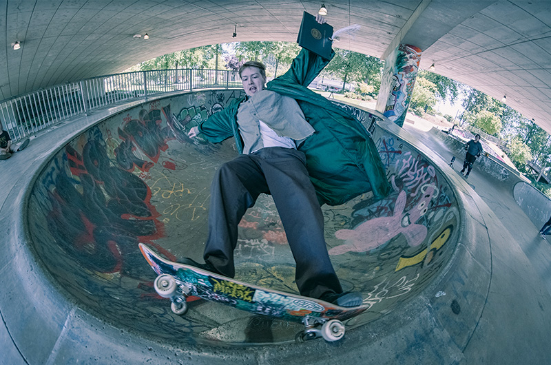 A student wearing a graduation gown and holding his diploma while skateboarding.