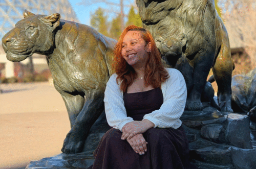 Kendra sits in front of a lion and lioness statue at the Omaha Henry Doorly Zoo with the Desert Dome in the background.