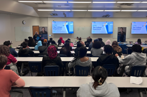 Picture of a full classroom with students seated at tables in a lecture hall. Panelists and presenters are at the front of the room with two screens on the wall stating the event title, “The College Experience: A Panel Discussion”