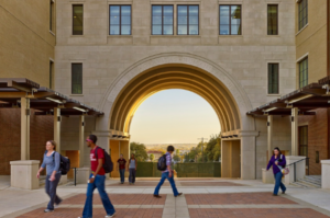 A scenic arch on Texas State University’s campus, with the sun setting behind it as students walk across a courtyard.