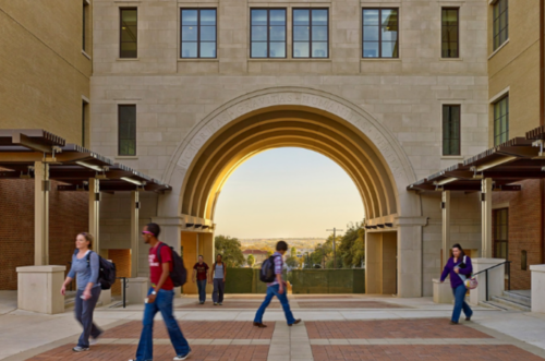 A scenic arch on Texas State University’s campus, with the sun setting behind it as students walk across a courtyard.