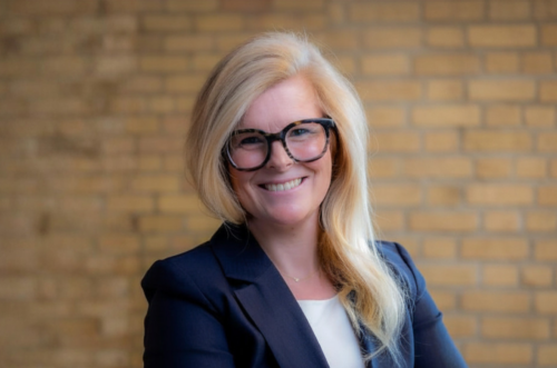 BG Tucker, executive director of College Possible Minnesota, folds her arms and smiles at the camera while wearing a navy blazer, with a modern, brick background behind her.
