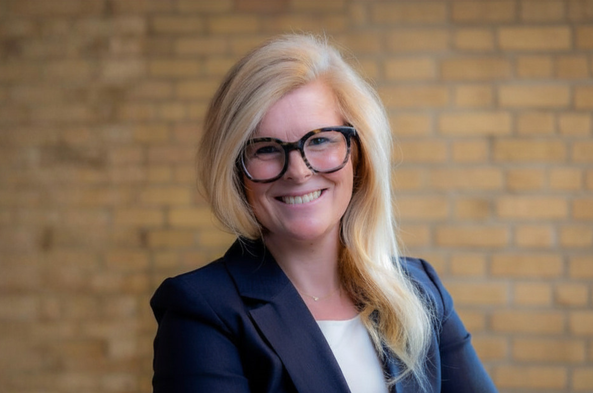 BG Tucker, executive director of College Possible Minnesota, folds her arms and smiles at the camera while wearing a navy blazer, with a modern, brick background behind her.