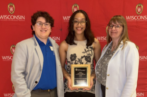 three college possible staff standing in front of UW-Madison background smiling holding a plaque