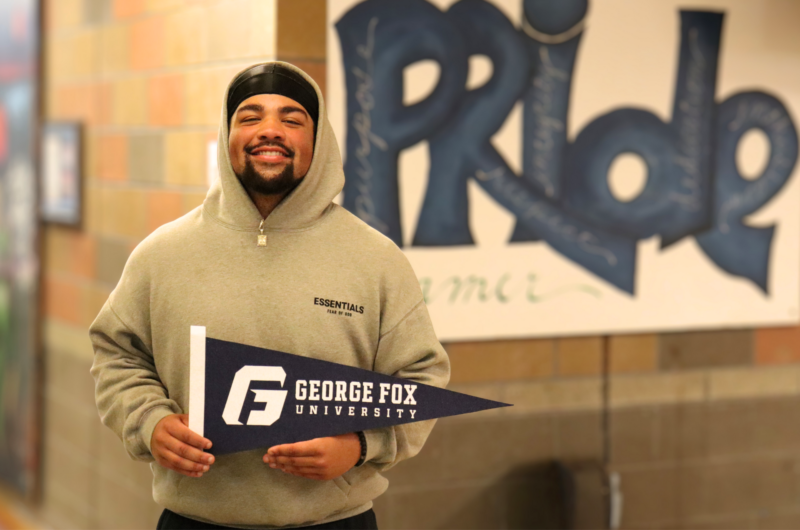 Dominic holds a George Fox University pennant while standing in front of a Todd Beamer High School poster that reads “Beamer Pride.”