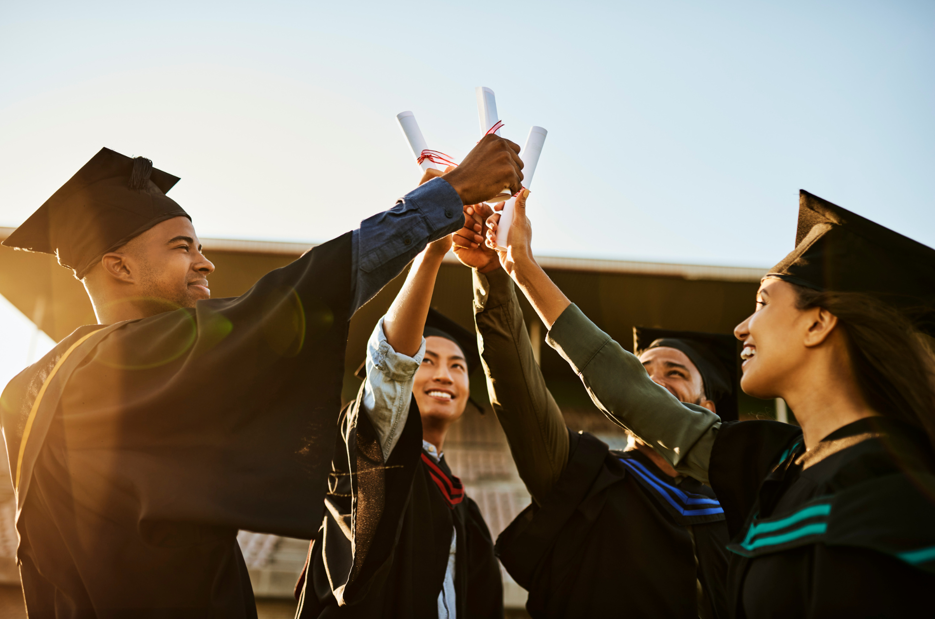 Four high school seniors stand in a circle, all wearing graduation caps and gowns, while celebrating their commencement diplomas