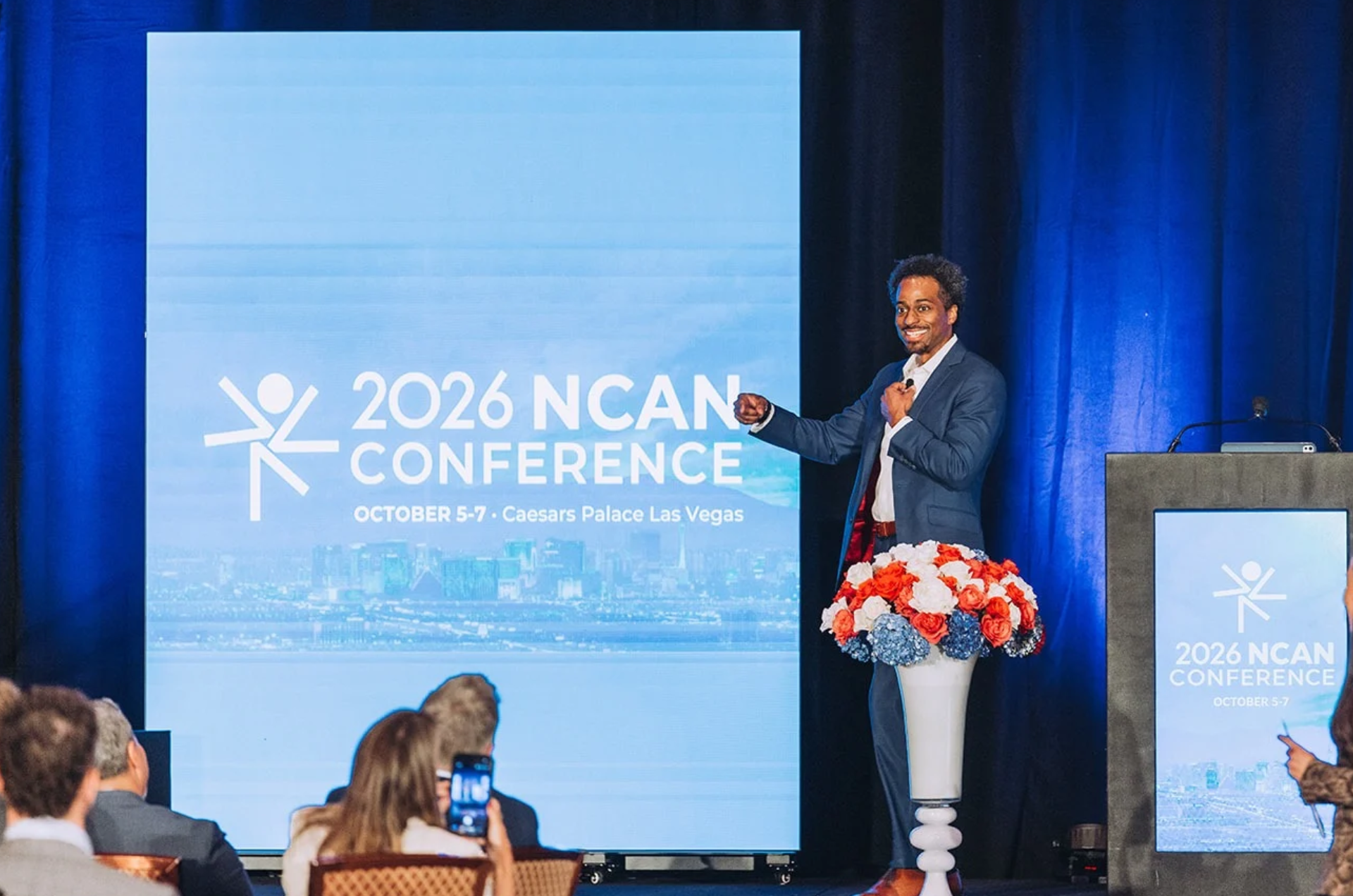 A lively Jesse Hendrix, pictured in a blue suit on a stage, speaks to a crowd of conference attendees seated at round tables.