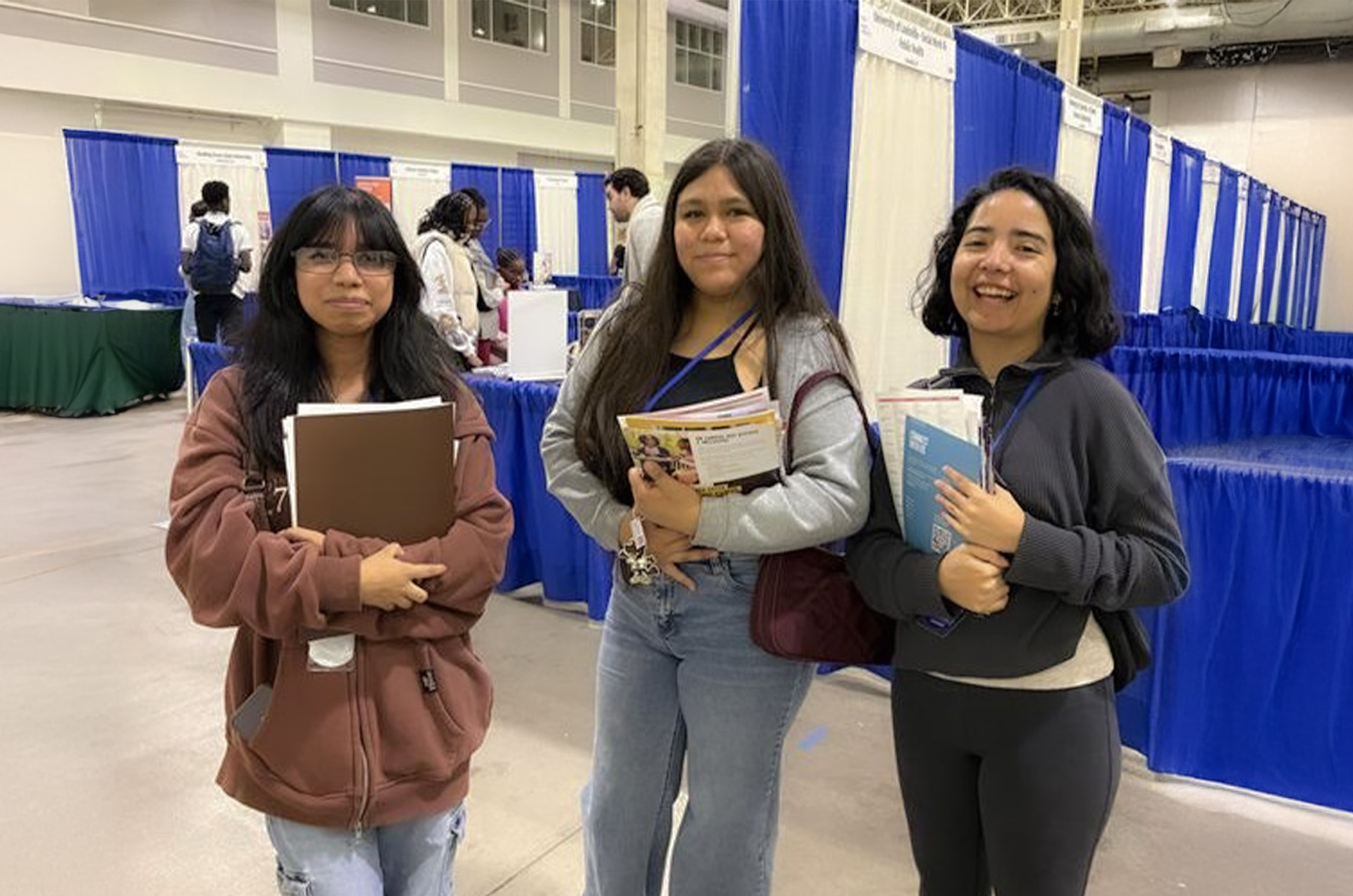 A group of students stand next to each other and hold up college brochures, smiling.